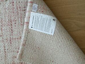 A woven rug is flipped over on a wooden floor, showing care labels and certification tags on the underside.