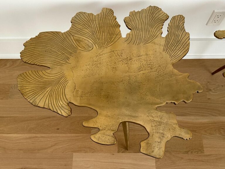 A gold, leaf-shaped metal table with textured, veined patterns on a wooden floor near a white wall.