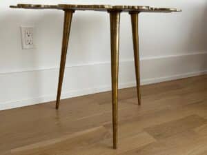 A small table with three gold legs and a scalloped edge top, standing on a wooden floor near a white wall outlet.