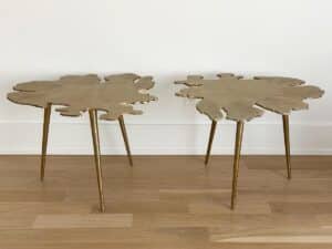 Two gold side tables with irregular, leaf-like tops and three slender legs each, on a wooden floor.
