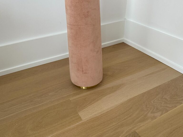 A pink cylindrical object with a gold base stands on light wood flooring in a corner with white walls.