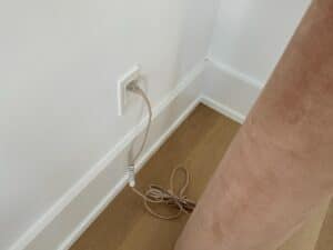 A beige cable plugged into a wall outlet near a light brown curtain and wooden floor.