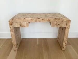 A light wood burl desk with two drawers, standing against a white wall on a wooden floor.