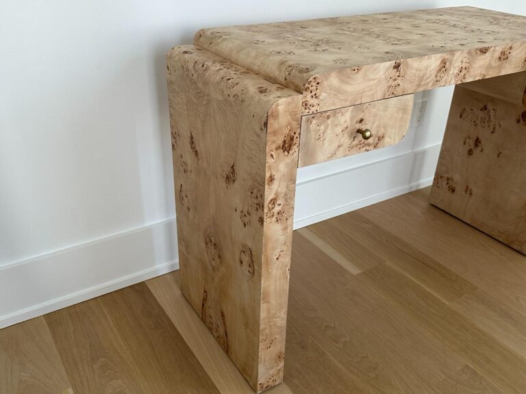 Modern wooden desk with rounded edges and a small drawer, placed on a light wood floor against a white wall.