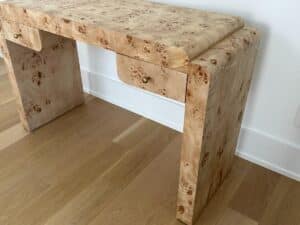 Light wood burl desk with rounded edges and two small drawers, placed on a light wood floor against a white wall.