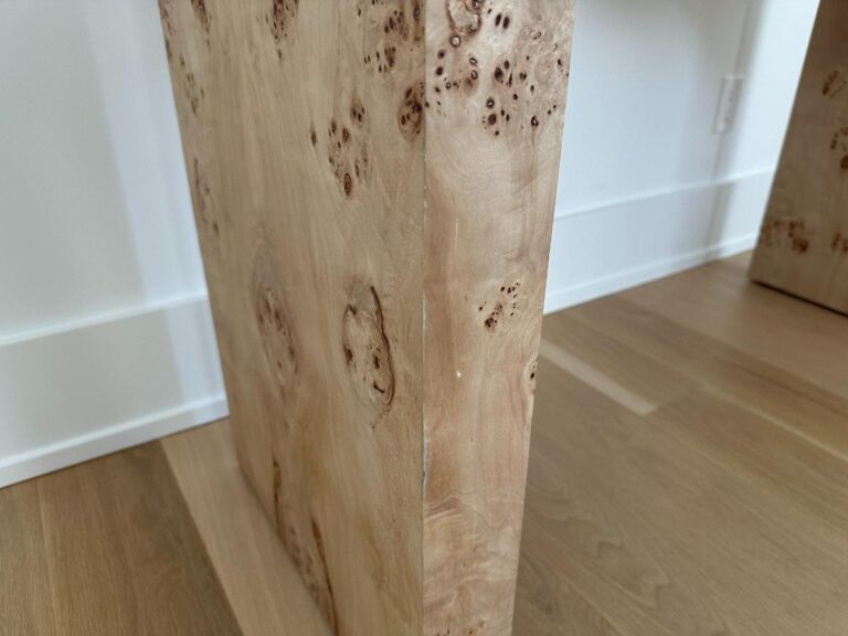 Close-up of a light wood table leg with a natural burl pattern, standing on a wood floor near a white wall.