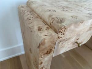 Close-up of a light wood desk with a burled texture and a small brass drawer pull, on a wood floor.