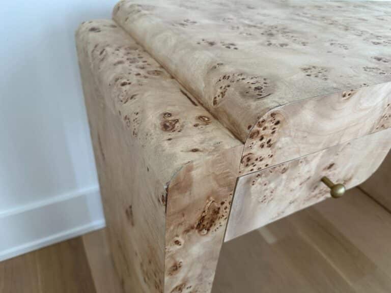 Close-up of a light wood desk with a burled texture and a small brass drawer pull, on a wood floor.