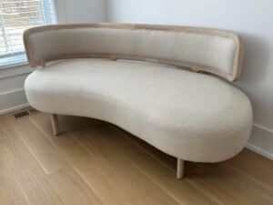 Modern beige curved sofa with wooden frame, placed near a window on light wood flooring.