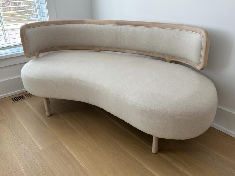 Modern beige curved sofa with wooden frame, placed near a window on light wood flooring.