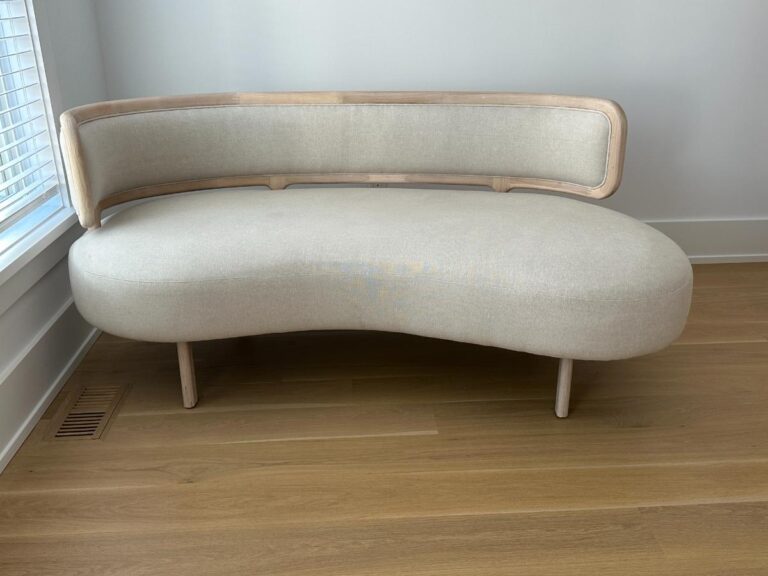 Curved, modern beige sofa with light wood frame on light wood floor near a window.