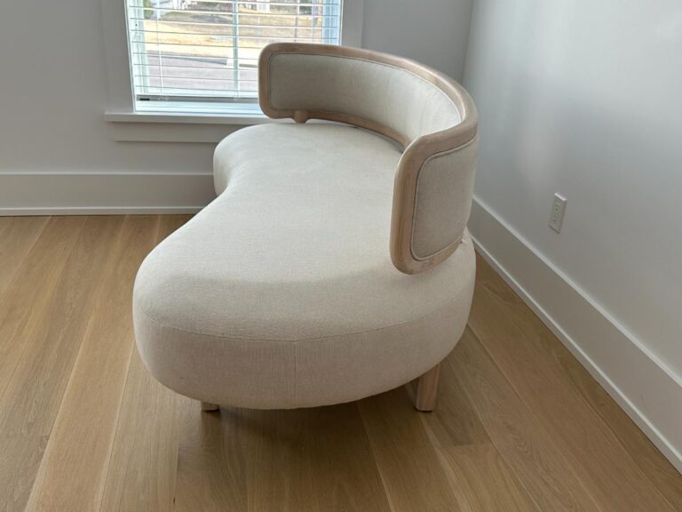 Curved, cream-colored loveseat with wooden trim, placed in a bright room with wood floors.