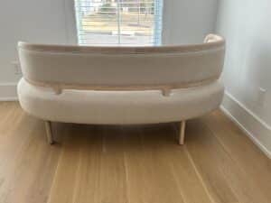 A beige curved sofa with wooden legs sits in front of a window on a light wood floor.