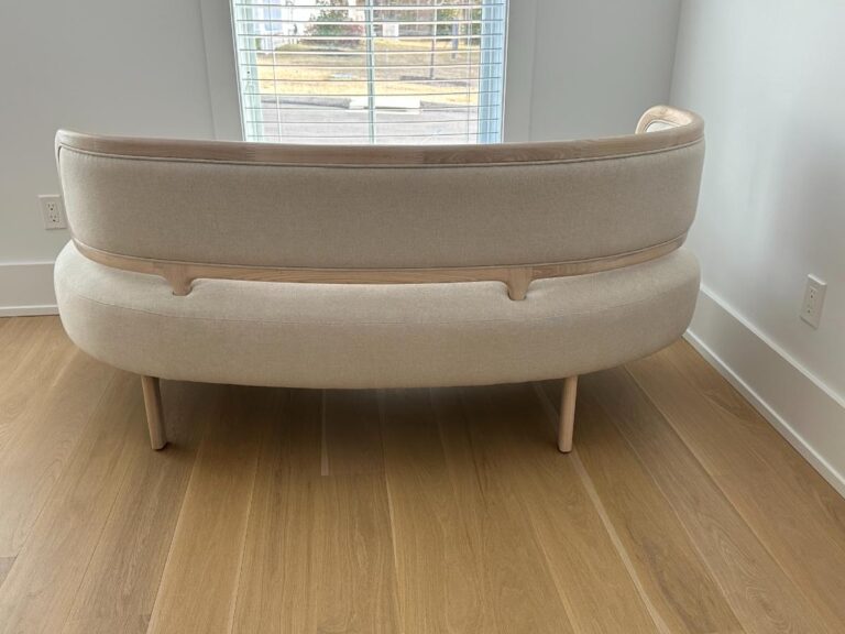 A beige curved sofa with wooden legs sits in front of a window on a light wood floor.