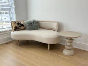 Curved cream sofa with two pillows beside a sculptural light wood side table in a bright room.