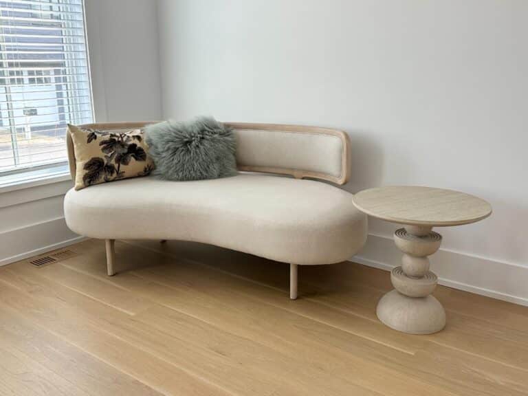 Curved cream sofa with two pillows beside a sculptural light wood side table in a bright room.