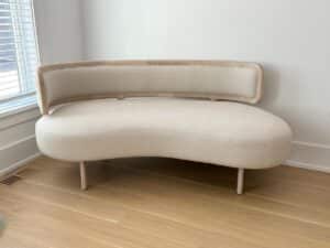 Curved beige sofa with wooden frame in a bright room with light wood floor and white walls.