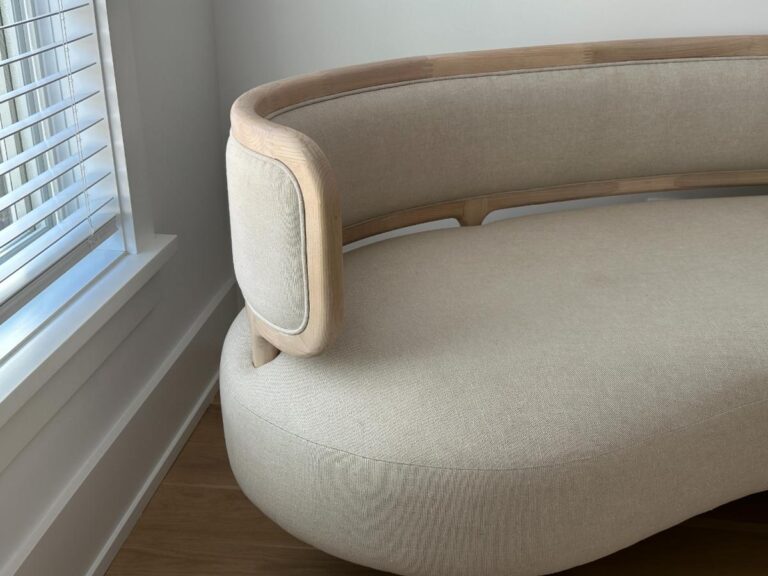 Curved beige sofa with light wooden frame near a window with white blinds in a bright room.
