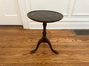 An Antique Side Table, small, round, and dark wood with three legs, sits on a wooden floor near a white paneled wall.