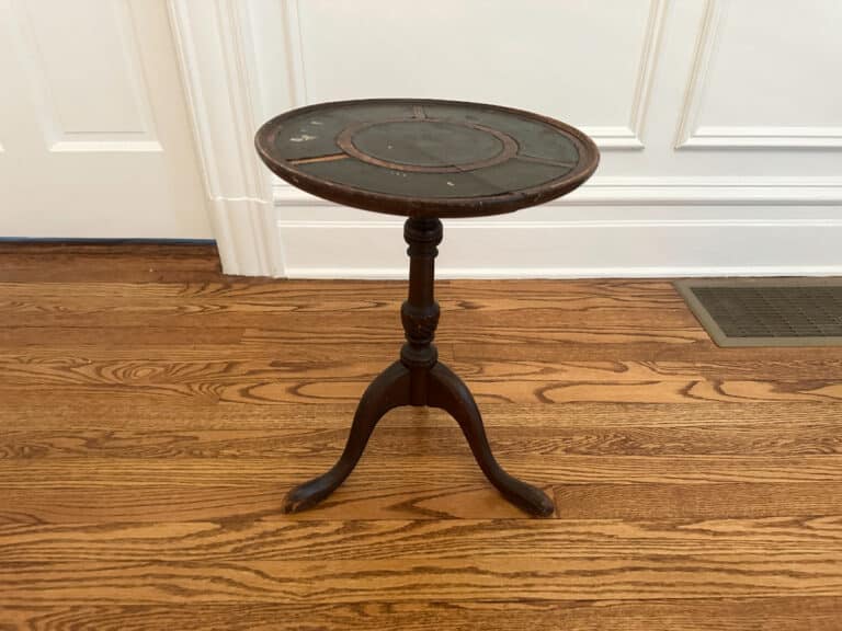 An Antique Side Table, small, round, and dark wood with three legs, sits on a wooden floor near a white paneled wall.