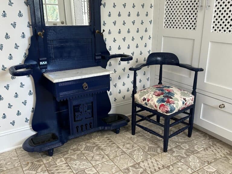 Blue vintage vanity with mirror and marble top, floral cushioned chair, and patterned wallpaper in a tiled room.