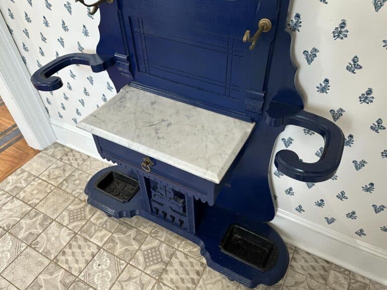 Blue wooden hall tree with a marble tabletop, hooks, and storage compartments, set against floral wallpaper.
