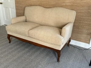 A beige upholstered loveseat with wooden legs, in a room with herringbone-patterned carpet and textured wallpaper.