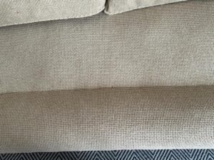 Close-up of a beige textured fabric sofa with a slightly darker tone at the bottom edge, on a patterned rug.
