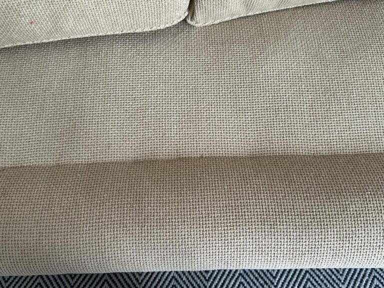 Close-up of a beige textured fabric sofa with a slightly darker tone at the bottom edge, on a patterned rug.