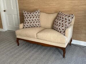Beige loveseat with patterned pillows against a textured wall and herringbone carpet.