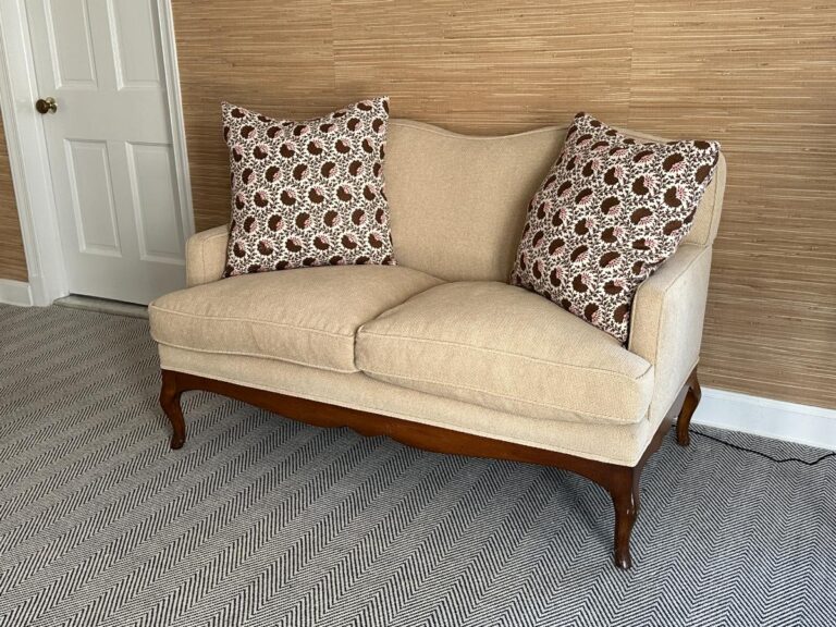Beige loveseat with patterned pillows against a textured wall and herringbone carpet.