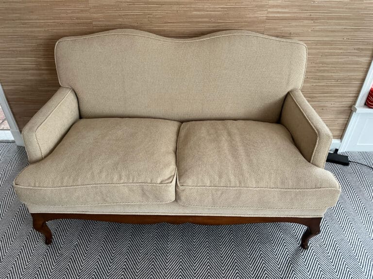 A Chenille Upholstered Settee with wooden legs on a patterned carpet in front of a textured wall.