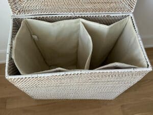 Open white wicker laundry basket with three cream-colored, divided compartments on a wooden floor.