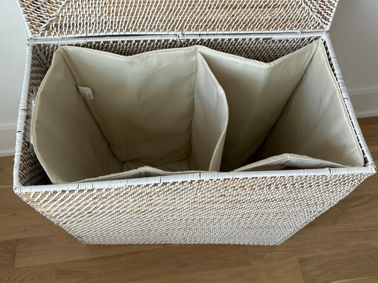 Open white wicker laundry basket with three cream-colored, divided compartments on a wooden floor.