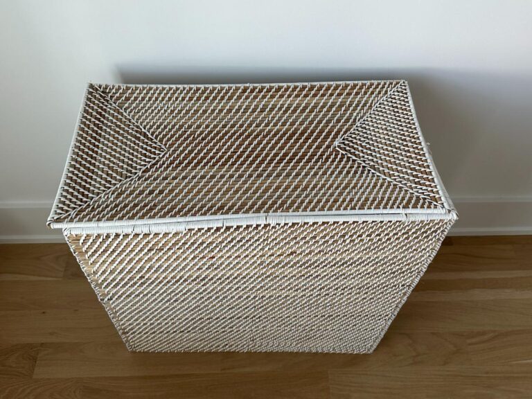 Rectangular white wicker laundry hamper with a closed lid, placed on a wooden floor against a white wall.