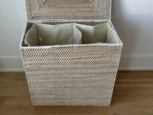 A white wicker laundry hamper with two beige fabric compartments, lid open, on a wooden floor.