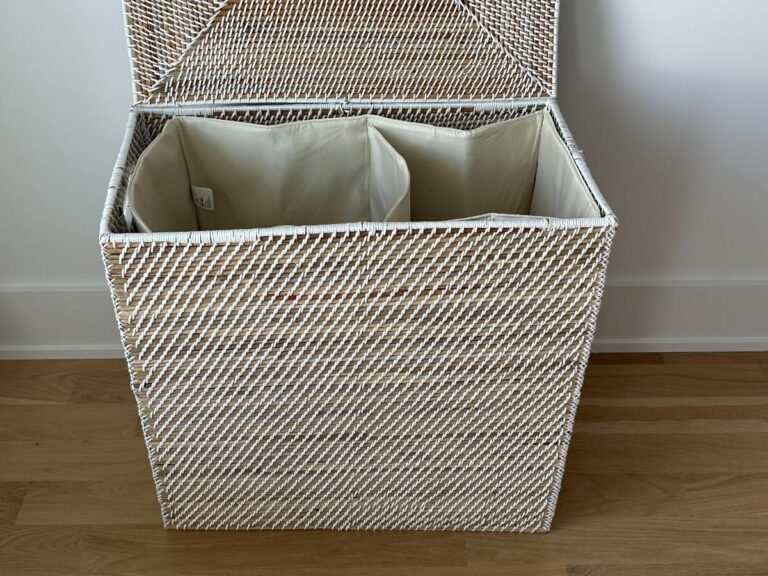 A white wicker laundry hamper with two beige fabric compartments, lid open, on a wooden floor.
