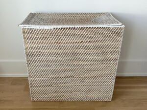 A rectangular wicker laundry basket with a lid, placed on a wooden floor against a white wall.