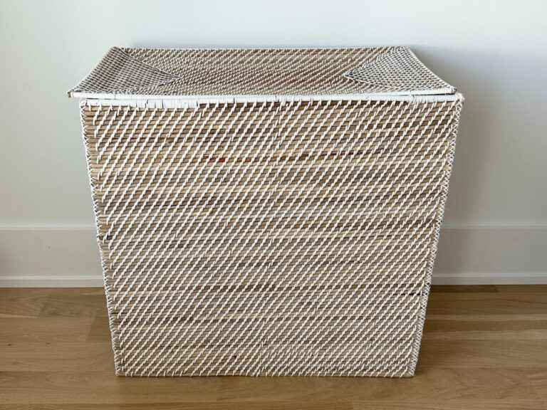 A rectangular wicker laundry basket with a lid, placed on a wooden floor against a white wall.