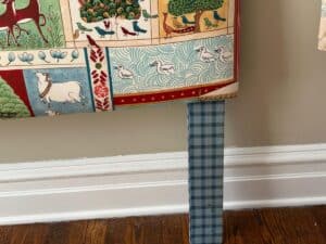 Colorful tapestry with animals and patterns draped over a blue gingham table leg, against a beige wall.