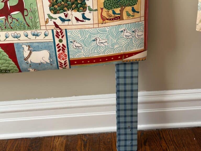 Colorful tapestry with animals and patterns draped over a blue gingham table leg, against a beige wall.