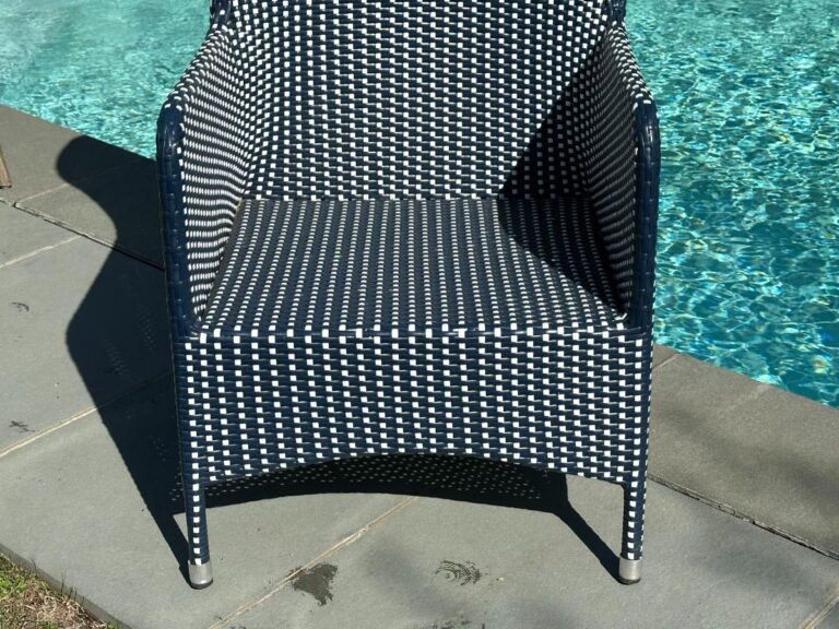 A black and white woven chair by a sunlit swimming pool on a stone patio.