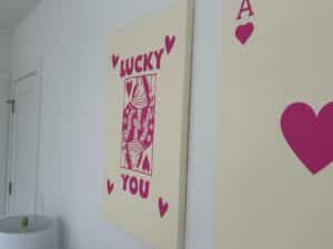 Beige wall art with a pink playing card design. Text reads "LUCKY YOU" with hearts around the card.