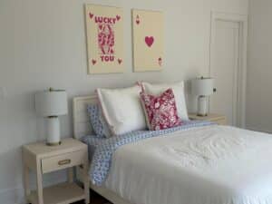 A neatly made bed with white and blue bedding, flanked by two lamps, and playful card art above the headboard.