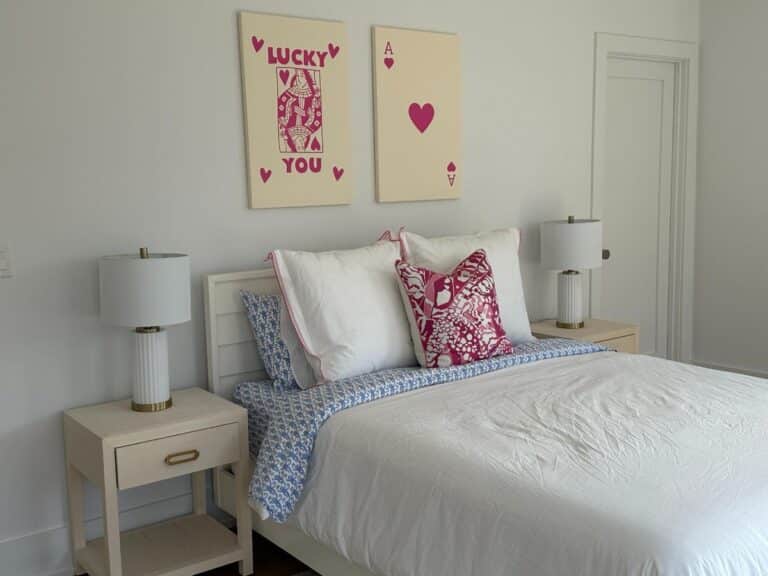 A neatly made bed with white and blue bedding, flanked by two lamps, and playful card art above the headboard.