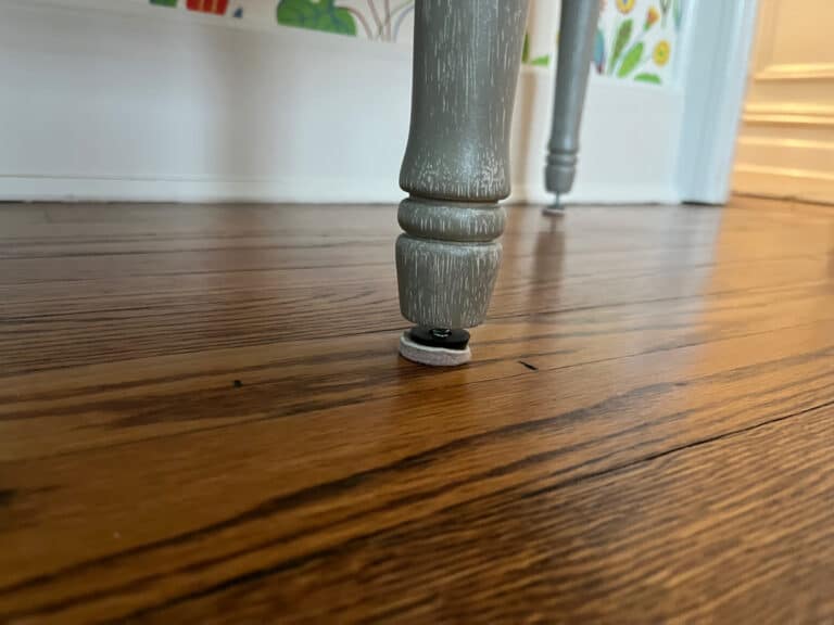 Close-up of a gray leg of the Demilune Tufted Bench with a protective pad on a wooden floor.