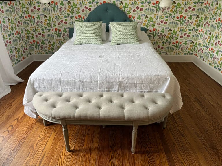 A neatly made bed with green pillows sits against floral wallpaper and a wooden floor, complemented by the Demilune Tufted Bench in beige placed in front.