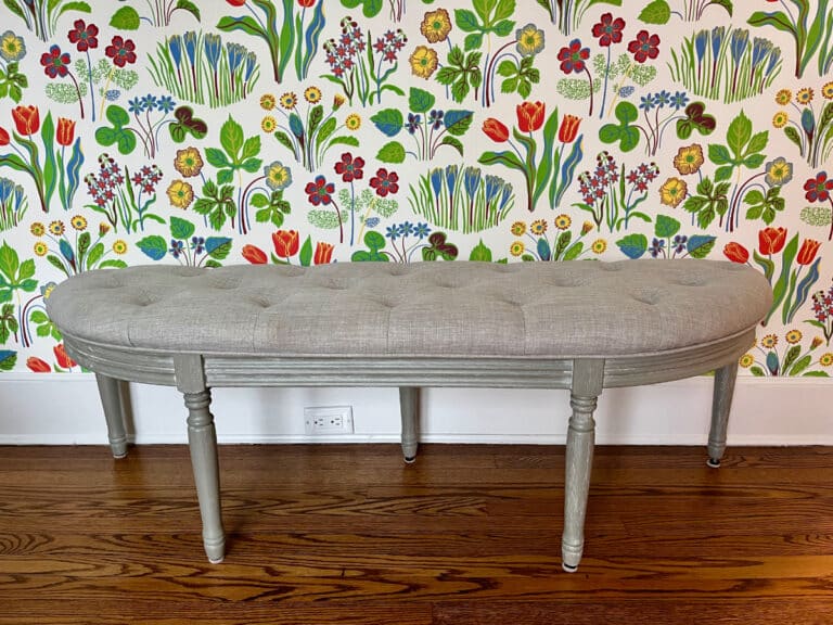 The Demilune Tufted Bench, with its gray upholstery, is set against vibrant floral wallpaper on a wooden floor.