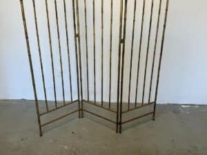 The Maison Bagues Gilt Bamboo Metal Folding Screen, featuring bamboo-style rods, stands on a plain concrete floor.