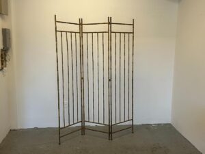 The Maison Bagues Gilt Bamboo Metal Folding Screen, featuring three panels, stands in an empty room with plain white walls and a gray floor.